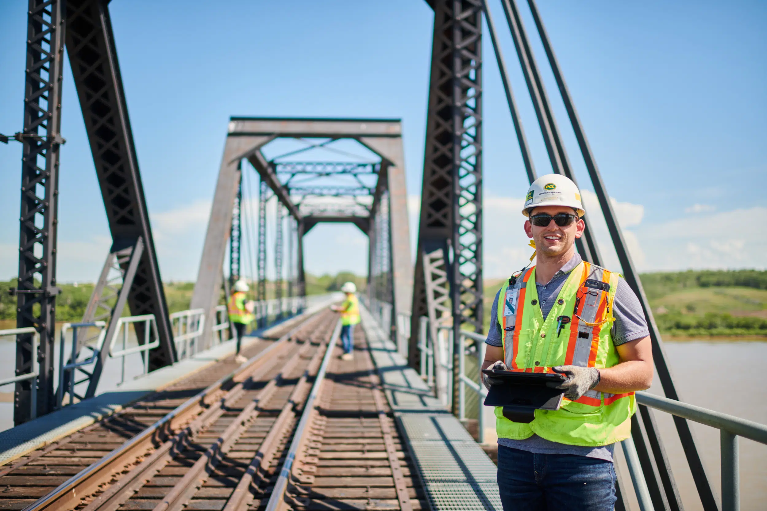 A construction worker stands on the top of a bridge with a tablet, surveying the area below.
