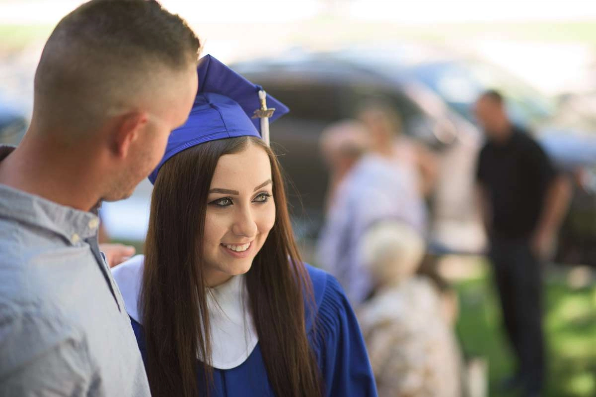 A medium close-up shot of a cdi student with a guest on graduation day. In this candid photograph, the smiling graduand greets guests prior to the ceremony. A shallow depth of field is used so that the student and guest are sharply in focus but the …