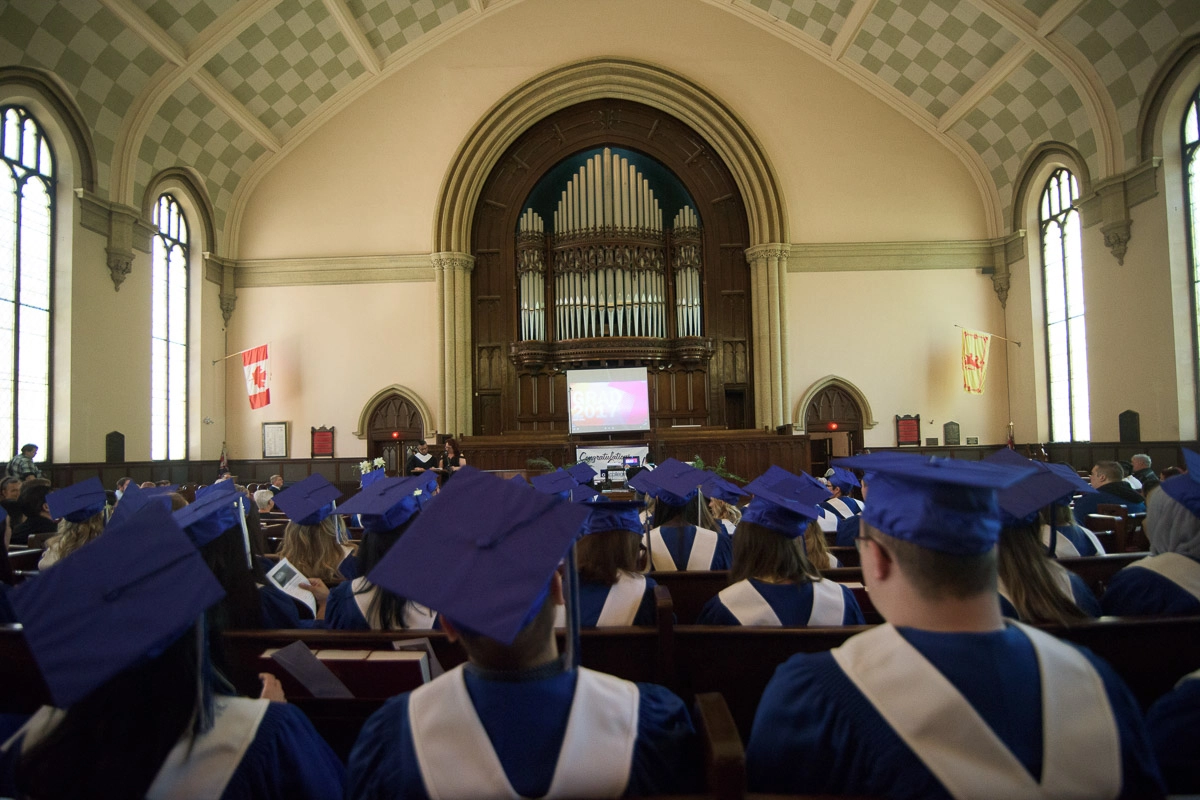 This photograph captures the rows of seated students and the area above them reaching up to the high vaulted ceiling. This perspective emphasizes the architectural features of the space, and provides a sense of its size and the number of people in t…