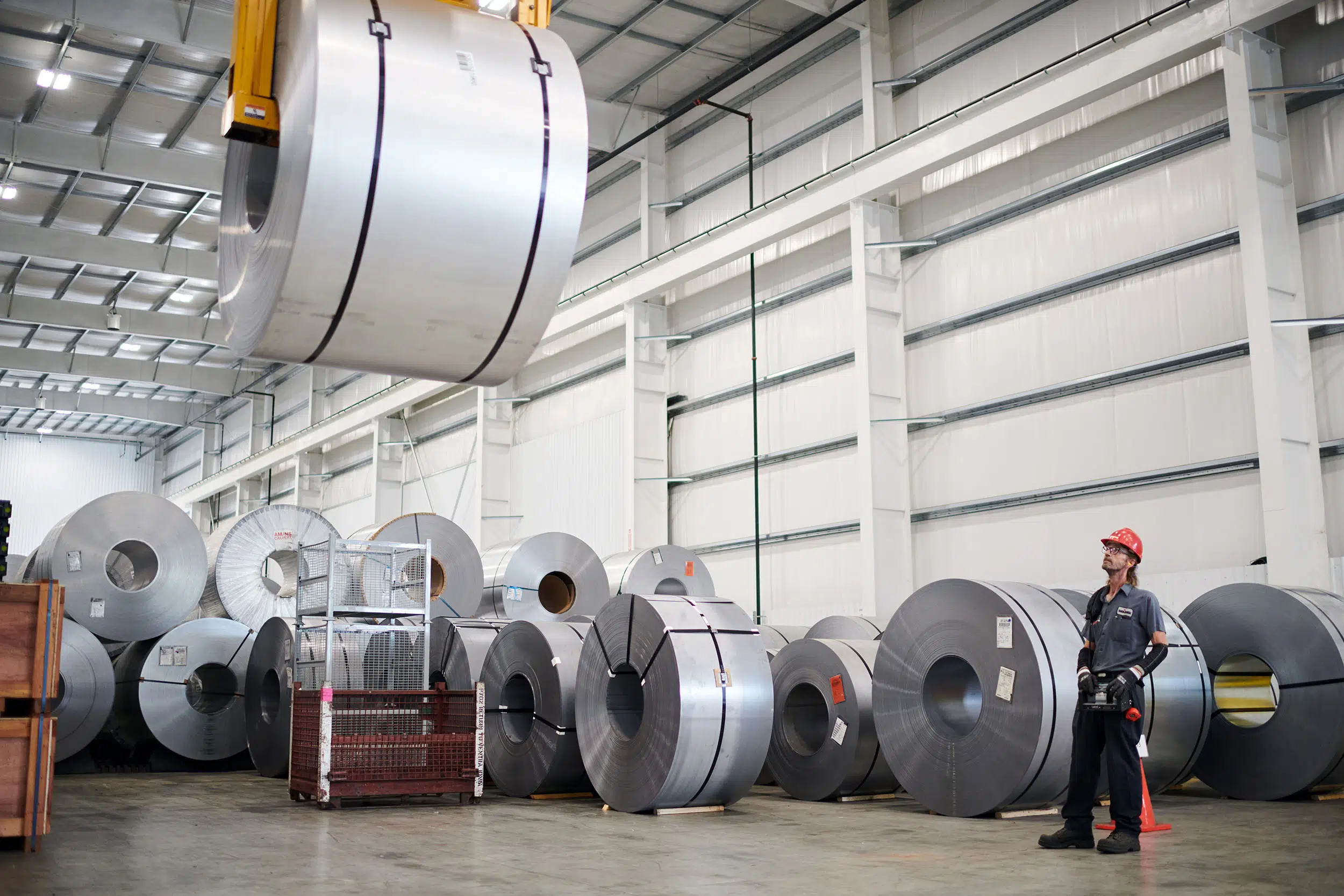 A worker uses a crane to move a large roll of metal.
